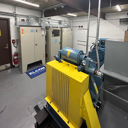 brightly lit machine room with updated yellow and blue machinery and two grey electrical cabinets on a white brick wall.