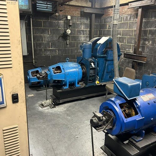 machine room with two large blue motors and other machinery, against a grey brick wall.