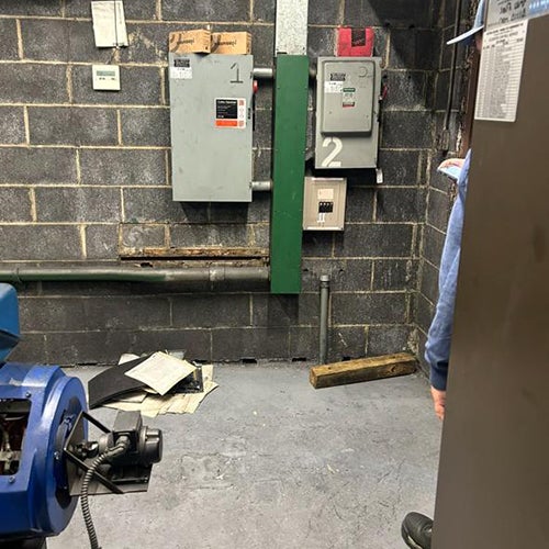 machine room with two large blue motors, exposed pipes, and electrical panels on a grey brick wall.