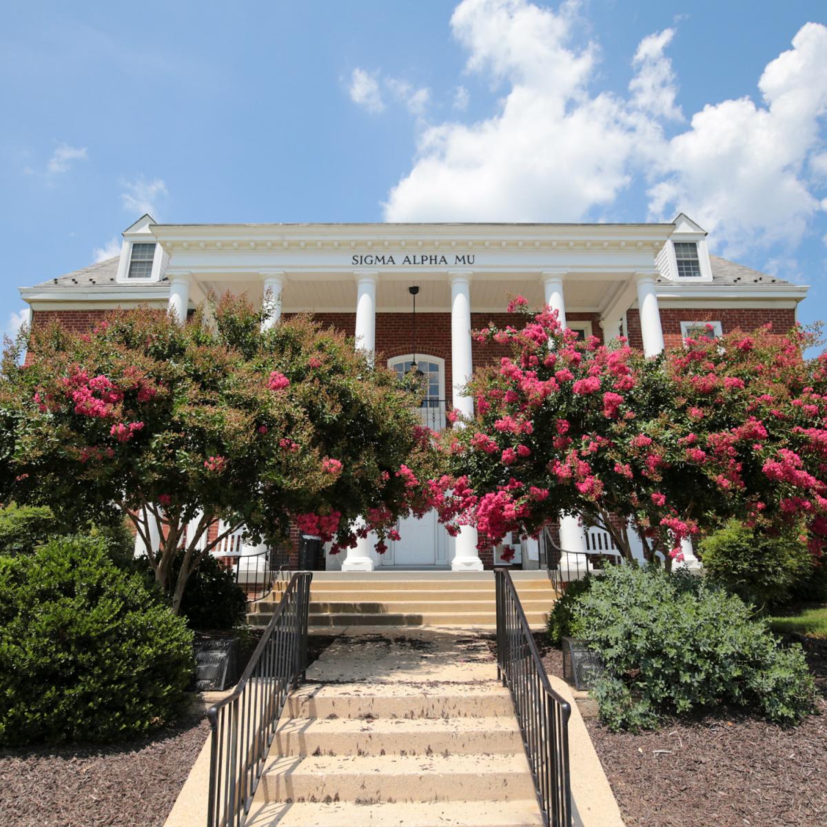 Brick house at 2 Fraternity Row with white columns and surrounding greenery and pink flowers