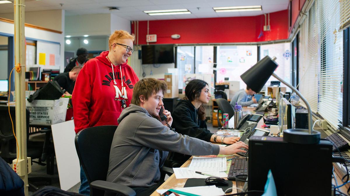 A staff member in a Maryland hoodie smiles while a student at a desk talks on an office phone and another types on a keyboard in the Service Center.