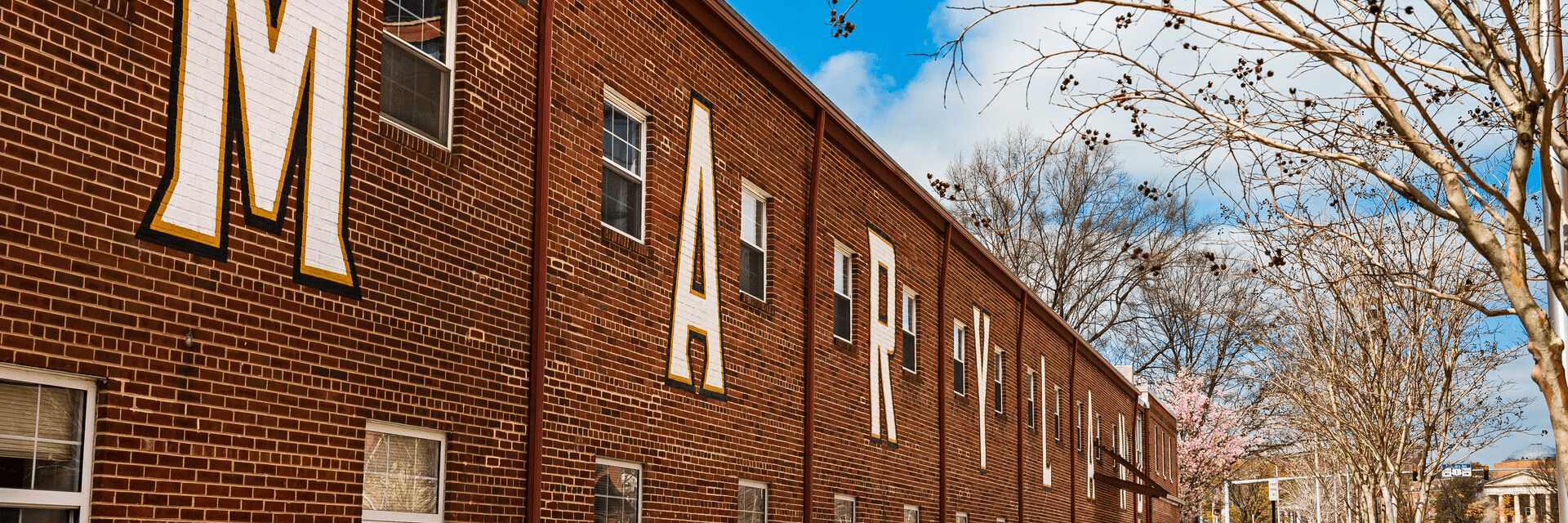 "Maryland" painted on the brick exterior of the Service building next to The Hotel against a blue sky with trees and blossoms.