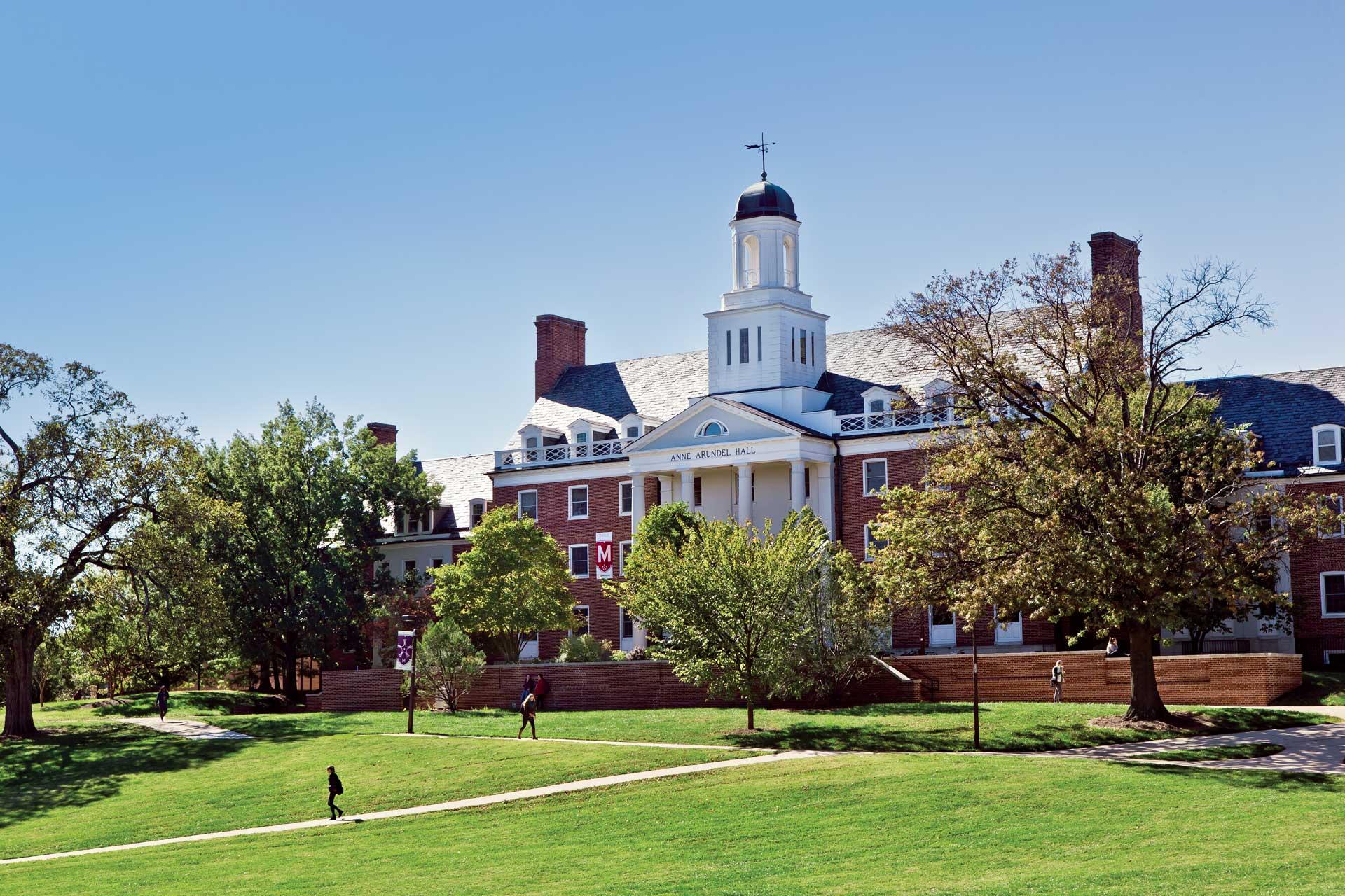 View of Anne Arundel Hall with students walking along the sidewalk in the foreground, with fall foliage.