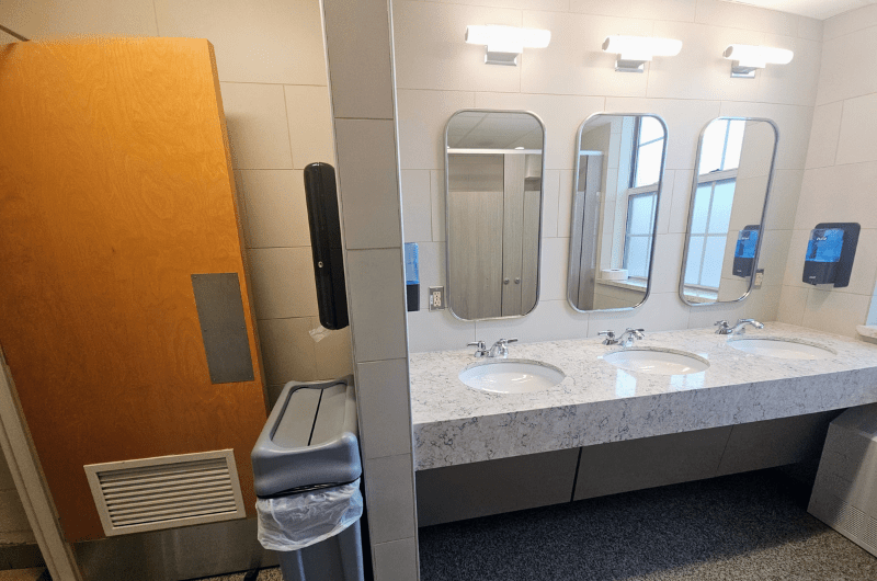 A newly renovated restroom sink area with three mirrors, sleek white counters, and touchless soap/paper towel dispensers.