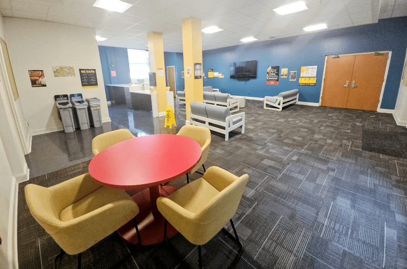 A brightly lit, newly renovated residence hall lounge featuring a red round table with four yellow chairs and blue accent walls.