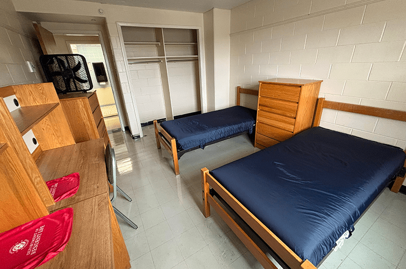 View of a standard double-occupancy residence hall room with two twin beds, two wooden dressers, and built-in desks.