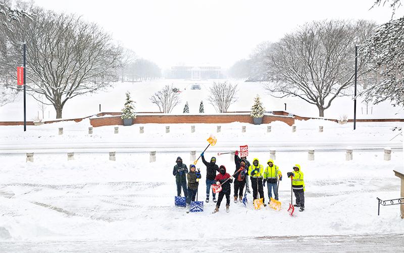 A group of snow shovelers, some in safety vests, pose and cheer on a snowy mall.