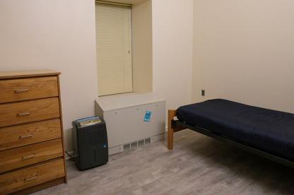 Portable dehumidifier set up on the floor of a residence hall room next to a bed and wooden dresser.